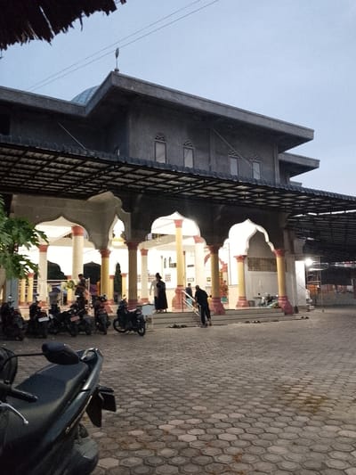 In Geulanggang Teungoh, Indonesia, an exterior shot captures a mosque with a gray, flat roof and a small dome, topped with a finial. The mosque features arched colonnades supporting a covered walkway, with yellow and red-accented pillars. Several motorcycles are parked in the paved courtyard. Inside the covered area, a few individuals are visible, some standing, one walking down a set of stairs, and another person dressed in black clothing is descending the stairs. The sky is overcast and the lighting suggests either dawn or dusk.