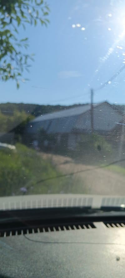The image shows a blurry view through a dirty car windshield of a rural landscape with a house and a dirt road. In the foreground, the dashboard of the car is visible, with a ribbed pattern on the edge. Sunlight is streaming in from the right, creating lens flare and glares on the windshield. In the background, a house with a light-colored roof is visible, partially obscured by trees. A dirt road leads towards the house. The sky is clear blue. The scene appears to be in a rural or suburban area, possibly the city of Кощаково, Russia, given the provided context. There are no people or animals visible in the image.