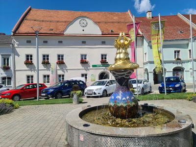 The image depicts a public square with a fountain in the foreground, located in Sankt Marein bei Neumarkt, Austria. The fountain is decorated with mosaic tiles and has water spraying upwards, with a golden statue of a seated figure on top. Behind the fountain, a street runs parallel to a row of buildings. Several cars are parked along the street, including a red sedan, a blue SUV, a white hatchback, and a blue compact car. Two flags are visible on poles to the right of the fountain, one pink and one yellow with text and graphics. The buildings are multi-story, with tiled roofs and visible windows. One building has a clock face on its facade. In the background, there is a clear blue sky with some white clouds. The overall scene suggests a sunny day in a town center.