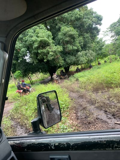 The scene depicts a rural or semi-rural area within the city of Akayar, Nigeria, observed from the interior of a vehicle. The foreground shows the vehicle's window frame, door panel, and a side-view mirror reflecting muddy ground and a person's boot. The midground features a heavily rutted, muddy dirt path flanked by dense green vegetation, including tall grass and undergrowth. In the background, several large, deciduous trees with full green canopies provide shade. Under and around these trees, a group of approximately 10-15 individuals are present, with at least eight motorcycles. Most motorcycles carry two occupants. One motorcycle on the left has a driver wearing a dark shirt and a red bandana, with a passenger carrying a backpack with red accents. Other individuals are dressed in dark or camouflage-patterned clothing. Some are seated on motorcycles under the trees, while others are positioned on a gentle, grassy slope to the right of the track. The sky above is overcast.