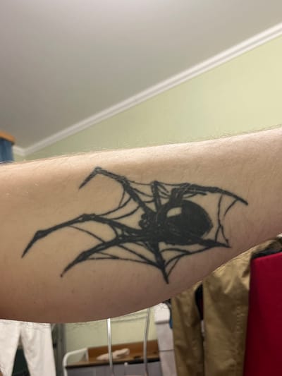 The image displays a close-up view of a tattoo on a person's forearm. The tattoo depicts a black spider in the center of a web, rendered in a stylized, graphic manner. The spider has a large, dark body and eight thin legs extending outwards. The web is abstract, with lines forming a rough circular shape around the spider. The skin of the forearm is visible, with fine hairs present. The background is out of focus, showing a pale green wall with white crown molding near the ceiling. The lighting suggests an indoor setting. The location of this image is not specified in the prompt.