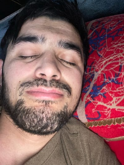 A bearded man sleeps with his eyes closed. His face is centered in the frame, slightly angled. He rests against a red pillow with blue and white patterns. A portion of a brown fabric covers his shoulder. His hair is dark and thick. The man’s lips are relaxed and slightly parted. The lighting is soft and somewhat uneven.