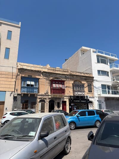 This image captures a street scene in Gżira, Malta, under a clear blue sky. The foreground is dominated by the front of a dusty silver car, its windshield bearing a parking ticket. To its right, the dark roof and side mirror of another car are visible. Behind these cars, a bright blue compact car is parked on the street.
Further back, a row of buildings forms the backdrop. The central building is an older, two-story structure with a weathered, pale yellow facade. It features two distinct enclosed balconies on the upper floor: one painted a vibrant blue with open windows, and another to its right painted red, also with open windows. Below these balconies, the ground floor of this building has several arched doorways and shop fronts. One prominent shop is labeled "ST. MATTHEW'S PHARMAÇY" with a black awning.
To the left of the central older building is a taller, modern, light-colored building with rectangular windows. To the right of the older building, another modern white building rises, featuring multiple levels with balconies and large windows, some with curved edges. Overhead, numerous electrical wires crisscross the scene, connecting the buildings. The overall impression is a typical urban street in Malta, showcasing a mix of architectural styles and parked vehicles.