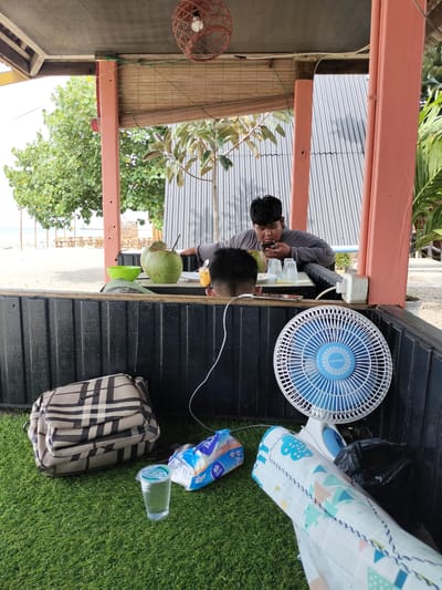 A young man in a grey long-sleeved shirt is seated at a table, looking at his phone and eating. In front of him are two coconuts with straws, a green bowl, and several plastic cups. Another person's head is visible in the foreground, facing away from the camera.
The scene is set outdoors under a rustic shelter with a woven bamboo roof and coral-colored poles. Behind the table, corrugated metal siding forms a wall. To the left, a beach and ocean are visible, with tables and chairs set up on the sand. Palm trees and other greenery surround the area.
On the grass-like ground in the foreground, there is a plaid handbag, a package of tissues, a plastic cup of water, a blue and white electric fan, and what appears to be a rolled-up blanket or mat with a colorful geometric pattern. A black trash bag is also present near the fan.
The lighting suggests it is daytime, likely sunny. The overall atmosphere appears relaxed and casual, possibly a beachside cafe or rest stop.
Visible text includes "Miyako" on the fan.