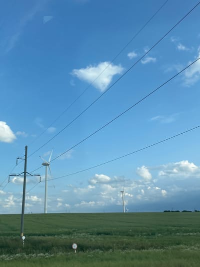 An outdoor shot captured from a slightly elevated perspective shows a vast, green, grassy field under a bright blue sky dotted with white clouds. In the mid-ground, several tall, white wind turbines stand on the horizon, with one prominent turbine to the left and others further in the distance. Power lines stretch across the sky from the left foreground, diagonally upwards and to the right. A solitary power pole is visible on the left side of the frame, positioned in front of the field and turbines. The field is mostly flat, with subtle undulations. The overall scene is serene and suggests a rural landscape. The image was taken in Ладзеніцкі сельскі Савет, Belarus.