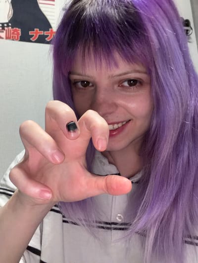 The image depicts a young female individual indoors in Lubnya, Russia. She possesses long, bright purple hair with bangs, exhibits a smile, and looks directly towards the viewer. Her right hand is positioned prominently in the foreground, curved into a claw-like gesture. The thumbnail of this hand displays a distinct solid black, square-shaped marking. She is dressed in a light-colored polo shirt with dark horizontal stripes near the collar. In the upper left portion of the background, a section of a poster featuring a monochrome illustration and visible Japanese text is affixed to a plain, light-colored wall.