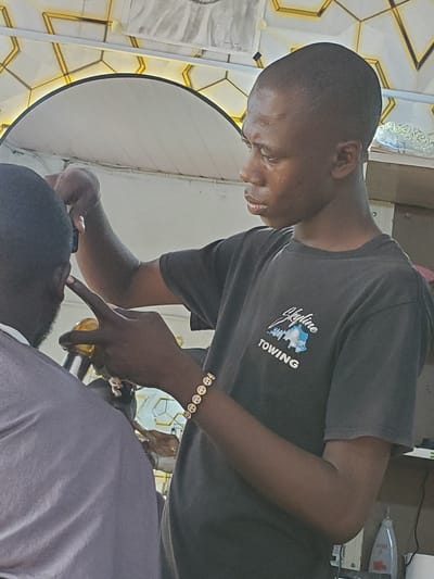 The image shows two people in what appears to be a barbershop in Ampan, Nigeria. One person, a barber, is leaning over the other person, who is seated or lying down, receiving a haircut or shave. The barber is wearing a dark t-shirt with a logo that reads "Skyline Towing". They are also wearing a bracelet with multiple smiley face charms. The barber's hands are focused on the client's face, with one finger touching the client's nose area. The scene is lit with bright lights, and a circular light diffuser is visible in the background. The wall behind the barber has a geometric pattern with golden accents, suggesting a modern or decorative interior. The overall mood is one of concentration and professional service. There are no clear indicators of the time of day or weather.
