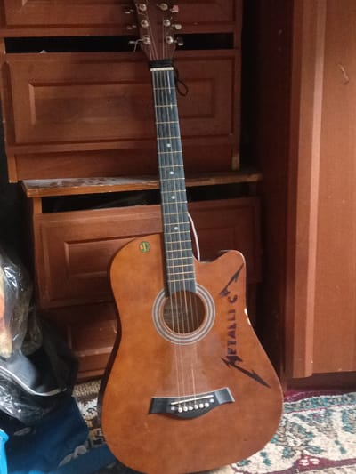 This is a medium shot, eye-level, full-body vertical image of an acoustic guitar leaning against a wooden cabinet. The guitar is a light brown color with a black pickguard and a soundhole with white and gray concentric circles. The guitar has six strings and a cutaway on the upper bout. A black logo resembling the Metallica band logo is drawn on the body of the guitar, with the text "METALLI C" written above it.
The background consists of a dark wooden cabinet with several drawers and shelves. To the left of the guitar, there's a black bag and a blue object partially visible, covered with plastic sheeting. A patterned rug is visible on the floor beneath the guitar.
The lighting is dim, suggesting an indoor setting. The image quality is moderate, with some graininess. No people are visible in the image. The overall mood is static and unremarkable, with the guitar being the sole focal point.
The text "METALLI C" and a stylized logo are visible on the guitar's body. A circular sticker with green and yellow colors is also visible on the upper bout of the guitar, but the text on it is illegible.