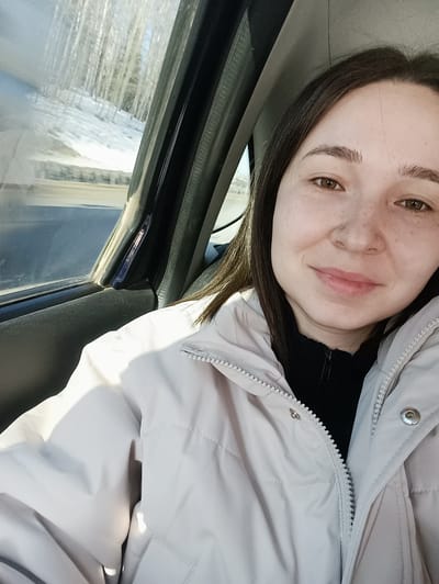 A selfie-style photograph depicts a single female subject within the passenger compartment of a vehicle, situated in Karashur, Russia. The woman, appearing to be a young adult, has dark brown hair, brown eyes, and visible freckles across her nose and cheeks. A small, spherical silver stud is present in her left nostril. Her lips are slightly parted in a faint smile. She is dressed in a light grey or beige puffer jacket, partially unzipped, revealing a black collared garment underneath. The jacket features a white zipper and snap buttons. To the left of the subject, through the vehicle's side window, a winter scene is visible. This includes a snow-covered embankment leading up to a dense stand of slender, leafless trees with light-colored bark. A blurred road surface and a metal guardrail are discernible at the base of the snowy incline. The exterior environment appears brightly illuminated by sunlight.