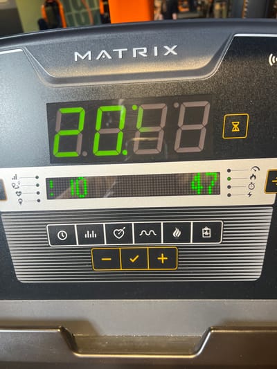 The image displays a close-up, head-on view of the digital control panel of a "MATRIX" branded exercise machine, situated within a fitness facility in Annœullin, France. The panel is constructed from dark gray and silver-colored plastic. The "MATRIX" logo is embossed on the upper section. A primary green seven-segment digital display shows "8.8:8.8," indicating a full segment illumination often seen in a test or default state. To the right of this display, a small amber hourglass icon is illuminated. Below, a dot-matrix display presents the number "10" on the left and "47" on the right in green pixels. To the immediate left of the "10" are vertical indicator lights and small icons depicting signal strength, a heart, an electrical plug, and a sound wave. To the right of the "47" are similar indicator lights and icons for a sound wave, an electrical plug, and a clock. Below these displays, a horizontal row contains five square buttons with white icons: a clock, a bar graph, a heart, a wavy line, and a flame. A sixth button depicting a battery icon is partially visible to the right. Beneath this row, three rectangular buttons are arranged horizontally within an orange border, marked with a minus sign, a checkmark, and a plus sign. The background is out of focus, revealing blurred orange and dark gym equipment, consistent with an indoor workout environment. No individuals are visible.