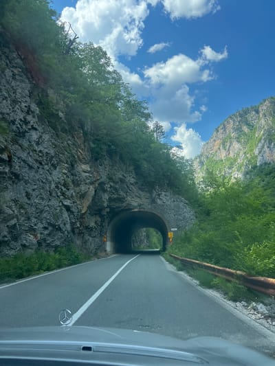 A front-facing, low-angle shot captures a road leading towards a tunnel carved into a rocky mountain face. The road is paved with a white line down the center and a Mercedes-Benz emblem is visible on the hood of a car in the foreground. Lush green trees and vegetation cover the surrounding cliffs and the tunnel entrance. The sky above is a vibrant blue with scattered white clouds. A rusted guardrail runs along the right side of the road. A yellow sign with black text and symbols is mounted on the wall of the tunnel. The location is Unknown, Montenegro.