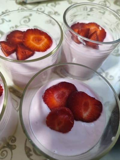 Four glasses of strawberry mousse topped with sliced strawberries are visible in the image. The glasses are clear and have a subtle pattern etched into them. The mousse is a pale pink color. The strawberries are bright red and sliced thinly, with some pieces cut into smaller, triangular shapes. The background is a light-colored, patterned tablecloth. The setting does not provide any information about the city of Cherneshchyna, Ukraine.