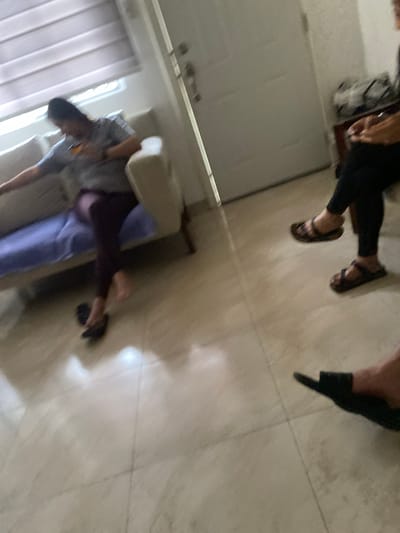 The scene appears to be indoors, likely a residential space given the presence of what looks like a bed or sofa, a window with curtains, and tile flooring.
People: There are at least two people visible, but both are partially obscured. One person appears to be seated, wearing dark pants and sandals. Another person is lying on what seems to be a bed or sofa, with a light-colored pillow or headrest.
Objects: Besides the furniture and window, there is a bag or purse near the seated person.
Actions: One person is seated while the other is resting on a bed or sofa.
Environment: The environment is indoors, likely a home or apartment.
There is no information given about the location of La Aurora, Ecuador, in the image.