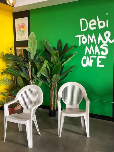 The image displays an interior scene of "Debi Tomar Mas Cafe," located at 3105 N Piedras St, El Paso, United States. The environment features a distinct wall color scheme: a bright yellow section on the left, separated by a narrow black vertical stripe from a vibrant green section that dominates the right. The ceiling is white, and the floor is a dark gray concrete.
Mounted on the black stripe and adjacent yellow wall is a framed chart resembling a "Coffee Taster's Flavor Wheel," characterized by multicolored segments. Below it, a smaller framed object is partially visible, showing text that includes "FOOD+WINE." On the green wall, large, hand-painted white capital letters spell out "Debi TOMAR MAS CAFE."
Two tall artificial banana plants with multiple faux trunks and large green leaves stand in black planters, positioned against the green wall. To the left, closer to the yellow wall, several smaller live green plants in reddish-brown pots are arranged. In the foreground, two identical, unoccupied white plastic chairs with slatted backrests and armrests are positioned on the concrete floor, facing slightly inwards. An exposed warm-toned light bulb hangs from the ceiling in the upper left quadrant. No individuals or active interactions are present in the scene.