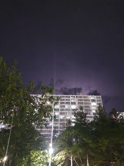 This is a night-time, low-angle, full-shot photograph of an apartment building in Gimhae-si, South Korea. The building is a multi-story structure with a white facade and numerous windows. Lights are visible in many of the windows, indicating occupancy. In the foreground, there are trees with green leaves, partially obscuring the lower portion of the building. Some streetlights or decorative lights are visible among the trees, casting a bright glow. The sky above is dark, with a purplish hue and some cloudy formations. There are no people or discernible actions visible in the scene. The overall impression is that of an urban residential area at night.
