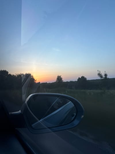 The image is a vertical, wide-angle shot capturing a sunset viewed from inside a vehicle. The primary subjects are the car's side mirror, the sky, and a silhouetted landscape. The mirror, positioned on the right side of the frame, reflects the interior of the car and the passing scenery. The sky displays a gradient of colors, transitioning from a pale blue at the top to hues of orange and yellow near the horizon, indicating it is either sunrise or sunset.
The setting appears to be a rural or semi-rural area, given the visible trees and the general darkness of the landscape, suggesting a drive through the countryside. The road is partially visible in the mirror's reflection. The overall mood is serene and contemplative, evoked by the warm sunset colors and the implied movement of the vehicle. The time of day is clearly indicated by the sunset. There are no people visible, and no text is present in the image. The location context is Dovhe, Ukraine.