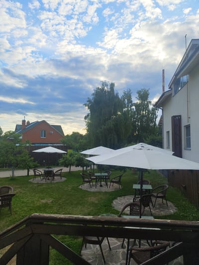 The image depicts an outdoor seating area with tables and chairs under large white umbrellas in a grassy yard. A brick house with a green roof is visible in the background, along with several trees and a wooden fence. The sky is cloudy with patches of blue. The scene appears to be a quiet, possibly residential or commercial establishment, in the city of Tsibino, Russia. There are no people or animals visible in the frame. The focus is on the architecture and the outdoor amenities.