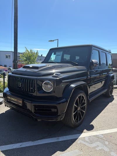 A black Brabus G-Wagon is parked on a paved lot in front of a building. The car is facing the viewer at a slight angle, with its front grill and hood prominently displayed. The grill features a large "B" logo. A license plate holder reading "ADS CONCEPT CAR" is attached to the front bumper. The lot is marked with white lines, and shadows from the car and other objects are cast on the ground. In the background, there is a building with windows, trees, and a utility pole. The sky is clear and blue. The scene appears to be in Mondercange, Luxembourg, as indicated by the text on the license plate holder.