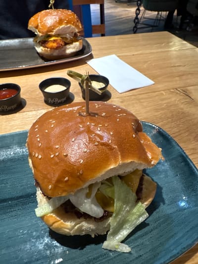 The image shows two partially eaten burgers served on separate rectangular plates, with three small containers of condiments on a wooden table. A small, folded white napkin is also present. One burger is on a dark-colored, textured plate, presented with condiments. The second burger is on a light blue plate. The setting appears to be a restaurant. The text "Le Burger" is printed on the condiment containers, suggesting the name of the restaurant. The city of Amstetten, Austria is not visually discernible from the image.