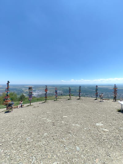 On a sunny day, a wide shot captures a scenic viewpoint in Goryachevodsky, Russia. The foreground is a gravel-covered plateau dotted with numerous signposts pointing in various directions, many adorned with colorful destination names. In the middle ground, a town stretches out below the plateau, nestled amongst rolling green hills and a distant hazy horizon. The sky is a vast, clear blue.

In the immediate foreground, on the left, a man with a backpack is crouched next to a signpost. Next to him, a woman is sitting on a wooden bench, looking towards the town. Further to the right, a cluster of signposts stands, with two people visible in the background. One person, a man, stands near a bench, while another person stands slightly further back. On the far right, a portion of a white picnic table is visible. The overall scene conveys a sense of tourism and exploration, with the signposts indicating a popular location for visitors to Goryachevodsky.