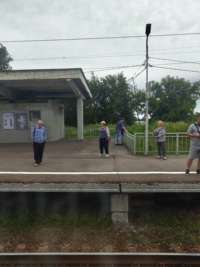 A group of five people are standing on a train platform in Konobeevo, Russia. The platform is covered by a shelter and surrounded by trees. The sky is overcast.
On the left, an older man wearing a blue shirt and dark pants stands with his hands in his pockets. To his right, a woman in a beige hat, blue top, and dark pants stands holding a bag. Further to the right, two men in blue shirts and jeans are leaning on a railing, one with a black bag over his shoulder. On the far right, a woman in a grey jacket and dark pants stands holding a black bag. The person closest to the camera on the right is a man in a grey t-shirt and shorts.