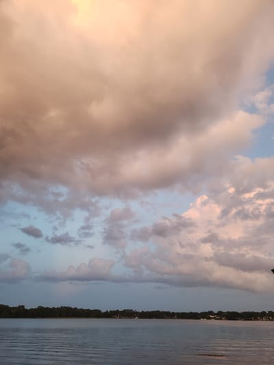 An image captures a wide expanse of the sky, dominated by voluminous clouds tinged with hues of peach, pink, and white, set against patches of pale blue. Below the sky, a calm body of water reflects the colors of the clouds, with subtle ripples disturbing its surface. Along the far bank of the water, a dense line of dark green trees forms a horizon, punctuated by a few scattered white structures that suggest the presence of buildings. The lighting indicates either dawn or dusk, casting a soft, diffused glow over the scene. This view is consistent with locations around lakes in Winter Park, Florida, given the typical landscape and atmospheric conditions common to the area. There are no people or animals visible in the frame.