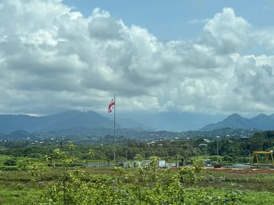 In Gvara, Georgia, a flag of Georgia flies on a flagpole. The flag is white with a red cross and four smaller red crosses in the quadrants. The flag is positioned in front of a backdrop of lush green hills and mountains. In the foreground, there are trees and vegetation. In the middle ground, there are some buildings, construction equipment, and a yellow gantry crane. The sky is partly cloudy with a mix of blue sky and white clouds. The overall scene is one of a rural landscape with signs of development.