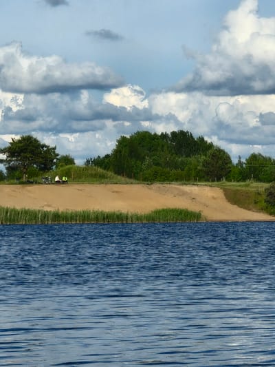 The image shows a scenic view of a body of water, likely a lake, with a sandy embankment and a line of trees in the background. The water has a rippled surface, reflecting the cloudy sky above. On the embankment, there are two people sitting, appearing to be engaged in conversation or resting. They are dressed casually, one in white and the other in green. Behind them, there are some trees and vegetation, suggesting a natural or park-like setting. The sky is partly cloudy with dramatic, dark clouds interspersed with lighter patches. The overall impression is one of a peaceful outdoor scene. It is not possible to determine the specific location in the city of Gargždai, Lithuania, from the image alone.