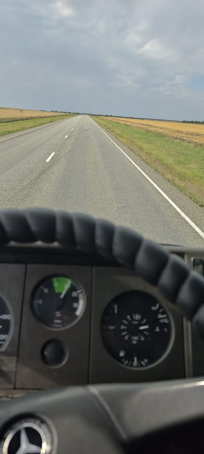 The image is a first-person perspective from inside a vehicle, looking out through the windshield. The vehicle is driving on a straight, paved road that extends into the distance. The road is flanked by flat, rural landscape with fields of dry grass or harvested crops on either side. The sky is overcast with gray clouds. In the foreground, the dashboard and steering wheel of the vehicle are visible. Several gauges are mounted on the dashboard. One gauge, on the left, shows a needle pointing to a green section, possibly indicating engine speed or temperature. Another gauge on the right appears to be a fuel gauge. The steering wheel has a thick, padded rim. The overall impression is of a journey through a vast, open countryside under a cloudy sky. The image is taken in Donskaya Balka, Russia.