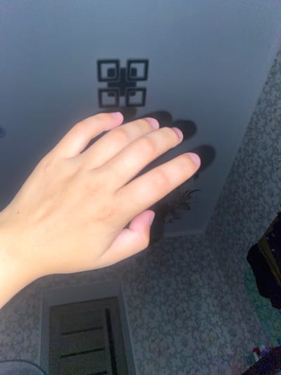 The image shows a hand with pale skin and light fingernails extended towards a ceiling. The ceiling is dark gray and features a geometric light fixture composed of four interconnected squares, forming a larger square shape. A shadow cast by the hand is visible on the ceiling, partially obscuring the light fixture. The background of the image is a wall with textured wallpaper, patterned with small, light-colored floral or abstract designs. To the right of the wall, part of a dark clothing item is visible. Below the wall and to the left, a doorway with a dark-colored door is present. The image does not provide any information about the city of Massy, Kyrgyzstan.