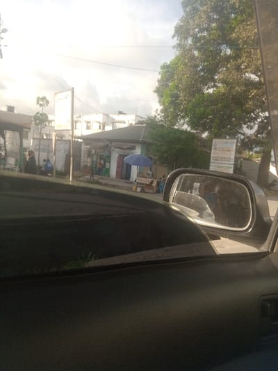 The image captures a daytime street scene in Magogoni ward, Kisauni Sub-county, Mombasa, Kenya, viewed from the front passenger side of a dark vehicle. The vehicle's dashboard and door panel occupy the foreground, with the passenger-side mirror reflecting part of the vehicle and additional scene elements on the right.
The environment consists of an urban street under a bright, partly cloudy sky. In the mid-ground, a light-colored, single-story building with a dark tiled roof operates as a commercial establishment, possibly a shop or cafe, identified by a partially visible sign above a blue market umbrella. The blue umbrella covers a roadside stall displaying items, possibly produce. Behind this, multiple multi-story, light-colored buildings are visible. To the far left, a structure with a dark canopy is present.
Several individuals are observed. One person stands near the blue umbrella and stall. To the left, a person wearing a blue shirt with the number "24" on the back is visible next to a motorcycle or scooter. Additional indistinct figures are present on the street. The side mirror reflects other people and parts of the street.
Key objects include:
* A tall, white rectangular sign on a pole to the left, with text obscured by brightness.
* A white sign on the right, partially obscured by a large tree, clearly displaying "ALL NATIONS CHOIR," "ENGLISH SERVICE 9:30 AM," "KISWAHILI SERVICE 11:30 AM," and "P.C.E.A KISIAUNI ACADEMY."
* Overhead utility lines.
* A patch of green grass is visible in the immediate foreground, just outside the vehicle.
The scene depicts typical street activity in a populated area.