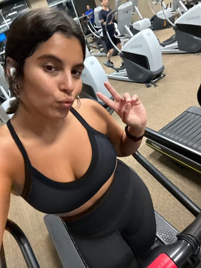 An image depicts a woman taking a selfie inside a gym, consistent with a fitness facility located in Plainsboro Township, United States. The primary subject is a woman with dark, wavy hair, wearing a black sports bra with mesh accents and matching black leggings. She has a white wireless earbud in her left ear and small hoop earrings. Her lips are puckered towards the camera, and her right hand is raised, forming a peace sign. A dark bracelet is visible on her right wrist. The background features several cardio machines, specifically elliptical trainers and a treadmill, which are in use by other individuals. At least two men are visible on elliptical machines. The gym floor is a light tan or beige color. In the immediate foreground, part of a black treadmill's console and a red emergency stop button are visible.