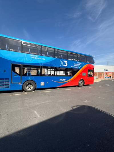 A blue and red "more" X3 double-decker bus is depicted parked on an asphalt surface under a clear blue sky with scattered cirrus clouds. The bus's side livery features "coast to cathedral city" with a skyline graphic, and lists route destinations including Salisbury, Fordingbridge, Ringwood, and Bournemouth, identifying the location as Salisbury, United Kingdom, likely at a bus station or depot. "Part of GoAhead" is also visible on the lower side. Indistinct human figures are faintly visible through the windows of the lower deck. Yellow dashed lines are painted on the asphalt near the bus's front-mid wheel. In the background, a light-colored commercial building with a promotional sign featuring orange elements and a person is visible. A prominent, dark shadow of the bus is cast onto the foreground asphalt, extending from the vehicle.