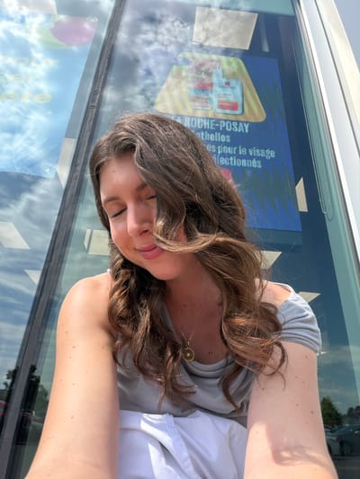 An outdoor, medium shot captures a young woman from the chest up, posing in front of a large glass building with a digital advertisement displayed on it. The woman has long, wavy brown hair that partially obscures her face. Her eyes are closed, and her lips are slightly pursed. She is wearing a light gray off-the-shoulder top and a gold necklace with a circular pendant. Her left arm is visible, showing several moles. The background shows a reflection of the sky and clouds on the glass, as well as trees and vehicles in the distance. The digital advertisement appears to be for La Roche-Posay, a skincare brand. This scene is located at a pharmacy in Longueuil, Canada, given the text on the advertisement.