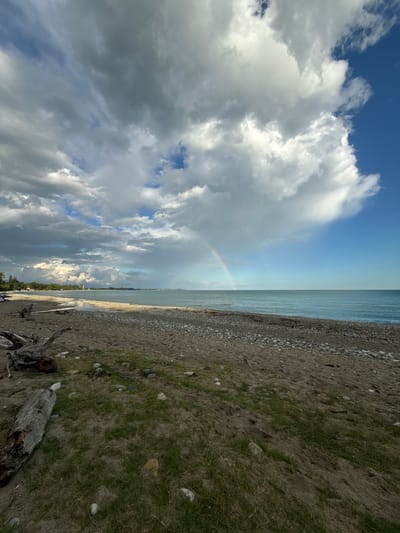 This media file depicts a wide, panoramic view of a natural landscape, likely a beach or shoreline, with a focus on the sky and water. The composition is split into distinct sections: the sky dominates the left side, filled with dramatic, cumulus clouds that are illuminated by sunlight, creating a sense of depth and movement. A faint rainbow arcs across a section of the sky, indicating recent or ongoing rain.

The right side of the image is divided between the tranquil, rippling water of a large body of water, possibly a lake or sea, and the shoreline. The shoreline is a mix of sand and pebbles, with sparse patches of grass growing near the water's edge. Some driftwood and larger stones are scattered along the beach, adding texture to the scene. The horizon line is sharp and clear, separating the sky from the water.

The lighting suggests it is daytime, possibly late afternoon or early morning, given the dramatic cloud formations and the quality of light. The weather appears to be mixed, with sunshine breaking through after a period of rain, evidenced by the rainbow and the bright illumination of the clouds. There are no visible people or specific objects that would indicate human activity, besides the driftwood. The overall mood evoked is one of natural beauty, serenity, and the dynamic interplay of weather elements. There is no visible text in the image. The location context suggests Mgudzyrkhwa, Abkhazia, which is consistent with a coastal or lakeside setting.