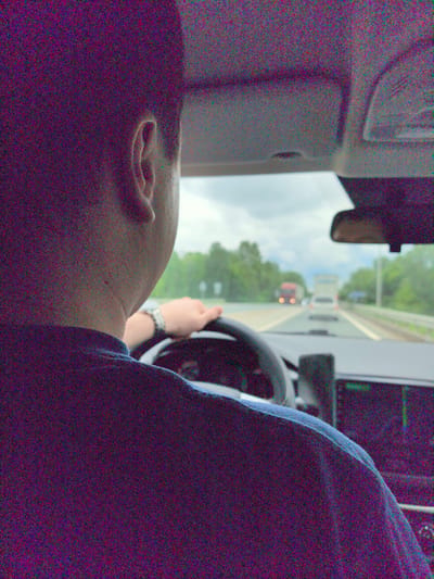 A man is driving a car on a highway. The driver is seen from the back, showing his head, ear, and upper body wearing a dark blue shirt. His hand is on the steering wheel. In the distance, there are other vehicles on the road, including a red truck and a white car. The environment outside the car includes trees and a cloudy sky. The car's interior elements are visible, such as the dashboard, rearview mirror, and headliner. No information points to the location of the city of Bol'shaya Sosnova, Russia.