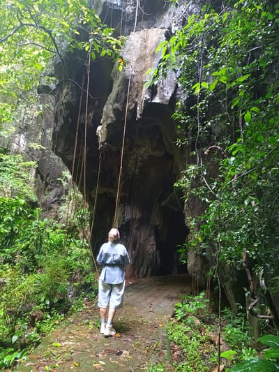 A woman stands at the entrance to a dark cave, located in Ban Pak Ra, Thailand. She has short blond hair, is wearing a light blue shirt, light blue shorts, white socks, and white shoes. The ground is covered with green vegetation. The cave opening is surrounded by large rocks and vines. The overall environment is lush and green.