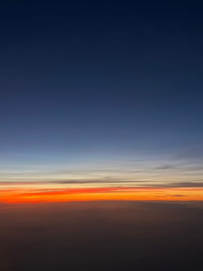 The image displays an aerial view of the sky at either sunrise or sunset. The upper portion of the frame is a deep, dark blue, transitioning downwards into a lighter blue, followed by subtle bands of grey and white clouds. Below these, a prominent, vibrant horizon line features intense orange and red hues, indicative of atmospheric scattering of light. The lowest portion of the image consists of a dense, dark cloud layer, appearing as an indistinct, undulating mass that extends across the bottom third of the frame. No ground features, man-made structures, people, or identifiable objects are present. The scene captures atmospheric conditions only. As an aerial view, no specific location within the city of Freising, Germany, can be identified from the visible details.