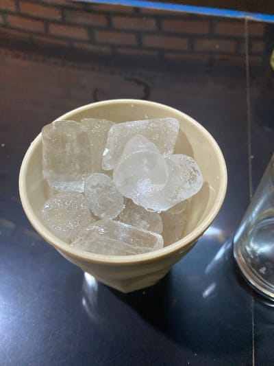 The image displays an off-white, faceted ceramic bowl filled with numerous irregularly shaped ice cubes, positioned centrally on a dark, reflective surface. The ice cubes are primarily clear with some exhibiting cloudy cores and show signs of melting, evidenced by moisture on their surfaces and small puddles of water on the surrounding dark surface. To the immediate right of the bowl, a portion of a clear glass, possibly a drinking glass or pitcher, is visible. In the blurred background, a brick pattern wall is partially discernible, topped by a horizontal blue stripe. The scene takes place in Đak Đoa, Vietnam. There are no individuals or discernible actions present in the frame.