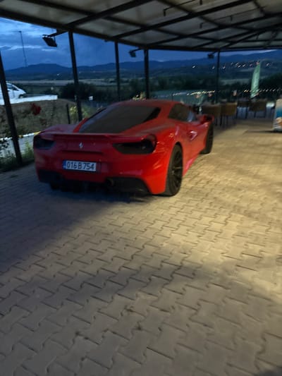 The image, rotated 90 degrees counter-clockwise, depicts a red car parked on a paved surface at what appears to be twilight or dawn. The car, a hatchback or small SUV, is shown from its rear quarter, revealing black multi-spoke wheels and dual exhaust pipes. A license plate displaying "016 B 754" is prominently visible on the back of the vehicle.
The paved ground consists of light-colored, interlocking concrete pavers laid in a herringbone pattern, transitioning from brighter illumination on the right to shadowed areas on the left. To the left of the car, a large glass barrier or window reflects a blue-grey sky and the silhouettes of distant mountains, indicating a scenic, possibly elevated, vantage point. A dark, angular structure, possibly part of an awning or building frame, partially covers the left side of the view through the glass. Faint lights are visible in the distant landscape, suggesting scattered buildings or infrastructure. The atmosphere is serene and quiet. Given the location context of Barakovo, Bulgaria, the mountainous backdrop is consistent with the region.