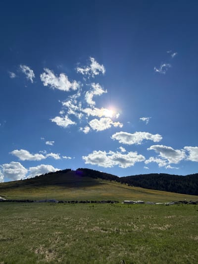 The image depicts a sunny day with scattered clouds over a rolling grassy hill. The sun is bright, peeking through some of the clouds, casting a glow across the sky. The hill is covered in green grass and some trees, particularly on the higher slopes. In the foreground, there is a grassy field with some rocks and what appears to be construction materials or debris near the base of the hill. The scene is an outdoor landscape, likely in a rural or semi-rural area. There are no people or animals visible in the image. The location is identified as Gela, Bulgaria, which is known for its mountainous terrain and natural beauty.