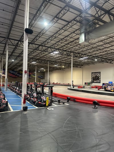 The image depicts the interior of an indoor go-karting facility in East Lake-Orient Park, United States of America. A series of go-karts are lined up in a blue-marked starting area, with additional karts visible further down the line. The track itself is gray with red safety barriers. A sign promoting "Birthday Parties" is visible in the distance. The ceiling is a high, exposed metal framework with lighting fixtures.