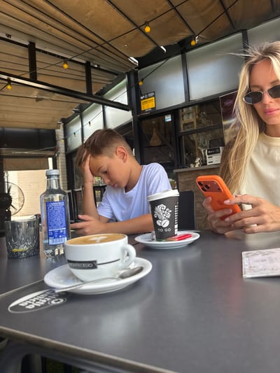 In the city of la Vila Joiosa, Spain, a boy sits at a table with his head in his hand, appearing distressed, while a woman to his right holds an orange iPhone and appears engaged with her phone. The table is set with a bottle of Cabreiroá water, a glass with ice and lemon, a coffee cup with latte art, a "To Go" coffee cup, and a small cookie. In the background, a fan is visible, along with a cafe setting featuring a bar and outdoor seating under a covered patio.