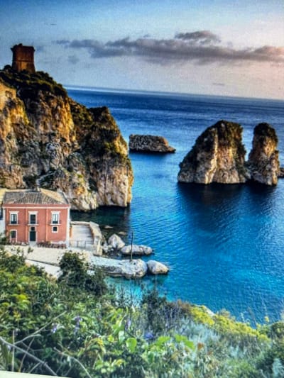 This media file depicts a coastal scene with rocky cliffs, the sea, and a building. The main subjects include several rocky outcrops jutting out into the vibrant blue water, a small brick building nestled into the cliff face, and lush green vegetation covering parts of the landscape. The scene is dominated by the natural elements of the sea and the rugged coastline, with the building adding a touch of human presence.
There is no discernible activity or event taking place; the image appears to be a still landscape. Notable details include the bright, clear blue of the sea, suggesting a sunny day or good weather. The architecture of the brick building, with its multiple windows, hints at a Mediterranean style. The rocky texture of the cliffs and the dense foliage contribute to the natural beauty of the location. The time of day is not explicitly clear, but the lighting suggests daylight, possibly mid-morning or afternoon.
No people are visible in the image, and there is no visible text. The overall impression is one of a serene and picturesque coastal environment, possibly in a Mediterranean setting, given the location context of Sesto Calende, Italy. The scene evokes a sense of tranquility and the beauty of nature combined with human habitation.