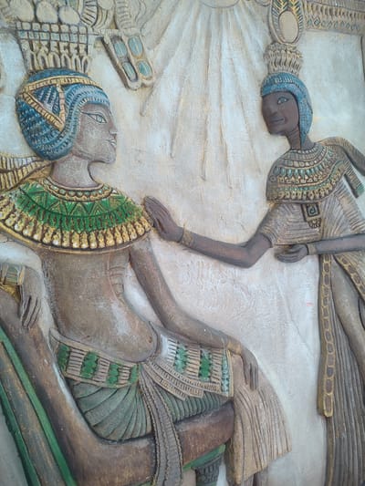 The image depicts a relief carving of an ancient Egyptian scene, likely Nefertiti and Akhenaten. Nefertiti sits on a throne. She is wearing a tall blue headress with a golden crown. She has a green and gold collar necklace, with a green and blue patterned belt. Akhenaten stands to her side with his hand on her shoulder. The scene is carved in beige and tan stone, with details highlighted in green, blue, and gold. The piece is found in Muang Pattaya, Thailand.