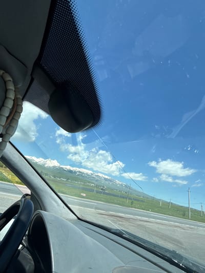 The image, taken from inside a vehicle looking through the windshield, shows a cracked windshield with a view of a landscape. Snow-capped mountains are visible in the distance, under a bright blue sky with scattered clouds. A road runs horizontally across the middle ground, with green fields and power lines flanking it. The interior of the car, including the dashboard, steering wheel, and a rearview mirror, are partially visible. There is a string of beads hanging from the interior of the car. The location is identified as Apnagyugh, Armenia.
