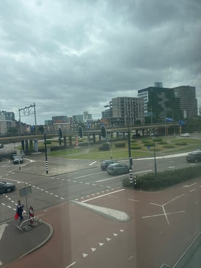 The image provides an elevated view of a complex urban intersection in Venlo, Netherlands, on an overcast day. The sky is uniformly grey with dense cloud cover, indicating no direct sunlight.
In the immediate foreground, a reddish-brown paved surface, likely a cycling or pedestrian path, occupies the bottom right, marked with white triangles and a distinctive white "Y" shape. Adjacent to this, a grey asphalt road section features dashed white lane markers. On the bottom left, a woman with light brown hair, dressed in a striped top and dark trousers, walks on a brick sidewalk, holding a red shopping bag. Next to her is an empty metal bicycle rack, beside a black and white striped post. A dark grey car is partially visible on the far left edge of the road.
The midground is dominated by a multi-lane road intersection. Numerous vehicles are present, including a light green compact car (possibly a VW Up!) moving left, a silver hatchback, a dark SUV, and a white compact car in the distance. Multiple sets of black traffic lights, some with white outlines, regulate traffic flow. An elevated concrete viaduct spans the intersection, carrying train tracks with overhead electrical lines and gantries. The support pillars of this viaduct are visibly adorned with colorful, abstract mural patterns. Several irregularly shaped grassy islands with low circular plantings and bushes are integrated into the road layout. A blue banner prominently displaying the word "Venlo" in white text is affixed beneath the viaduct on the right, confirming the city location.
The background consists of a diverse urban landscape. To the left, several lower-rise residential buildings with balconies are visible. In the center-right, a prominent dark modern building is largely covered by green vegetation, forming a vertical garden facade. Adjacent to it stands a taller, lighter-colored contemporary building characterized by multiple horizontal bands. Other urban structures, varying in height and style, recede into the distance under the heavy cloud cover.