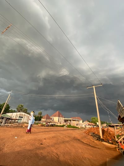 In Jere South, Nigeria, a woman in a blue head covering and red and white patterned attire walks along a dirt road, carrying a basket of items on her head. The sky is filled with dark, ominous storm clouds. In the background, several buildings with peaked roofs, some with multiple stories, are visible. Palm trees dot the landscape, and utility poles with electrical wires crisscross the scene. The ground is a reddish-brown dirt, with piles of construction materials and tires scattered near the roadside. The overall atmosphere suggests an approaching storm.
