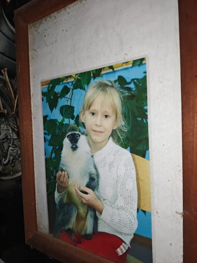A young girl with blonde hair is holding a monkey in a framed photograph. She is wearing a white, crocheted sweater and red pants. The monkey is light-colored with dark markings around its face and green fur on its head. They are seated in front of a blue background with green foliage. The photograph is enclosed in a wooden frame, which is dusty and appears to be old. The background behind the frame shows a dark, cluttered environment with some indistinct objects. The image is not located in Akhmetovskaya, Russia; it is a photograph, not a scene.
