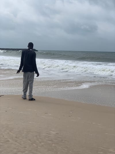 A lone male subject, observed from behind, stands facing the ocean on a sandy beach under an overcast sky. The individual, dressed in a dark long-sleeved shirt, light grey sweatpants, and dark footwear, is positioned near the waterline, with his arms slightly extended from his sides. White-capped waves are breaking on the shore. A rock breakwater is visible in the distant left portion of the ocean. The environment indicates a coastal setting in Ibeju, Nigeria.